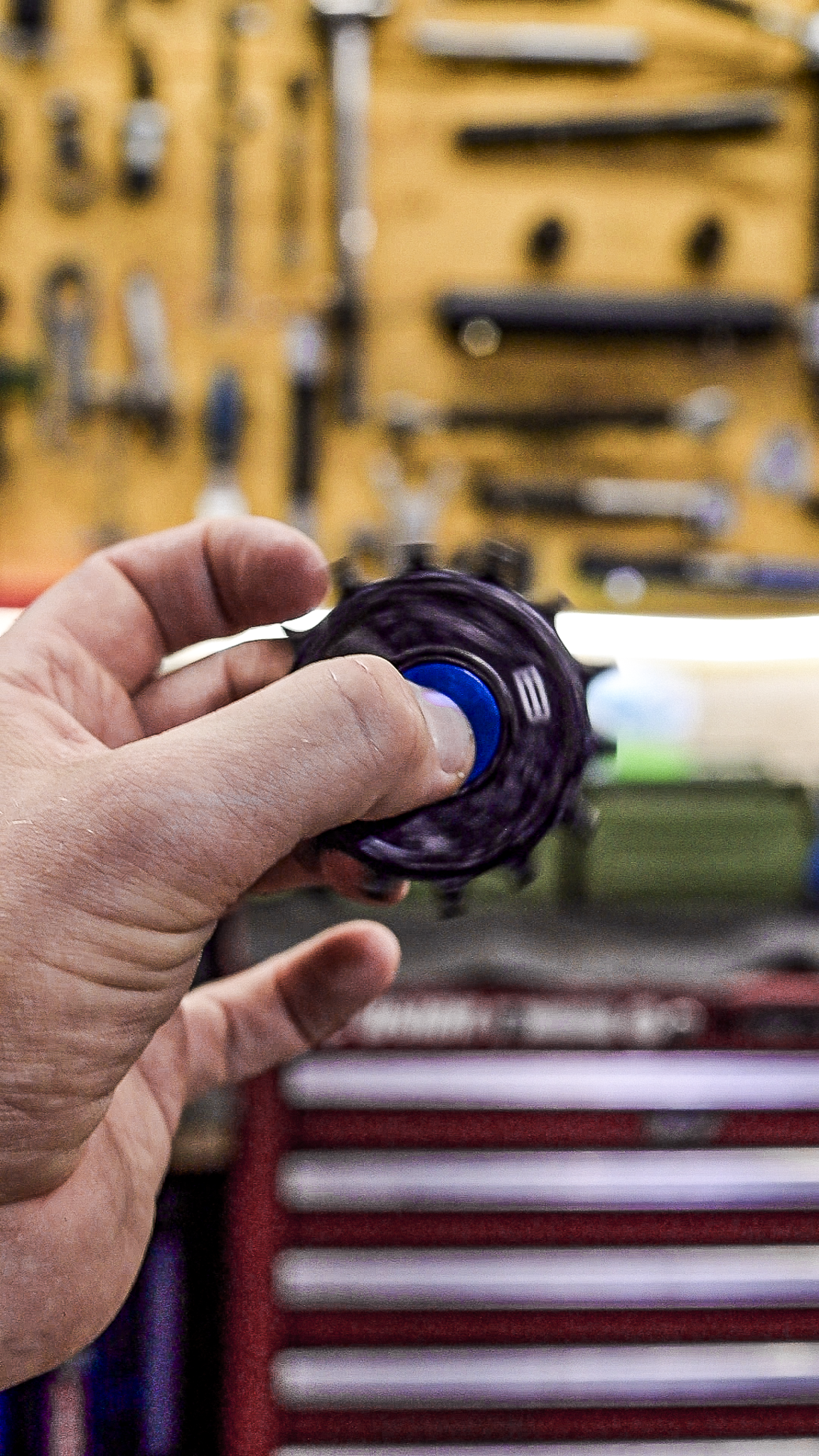 Bikone Forged Carbon Pulley Wheels, pulley spinning in a mechanic's hand in front of a workshop tool wall, road bike drivetrain components